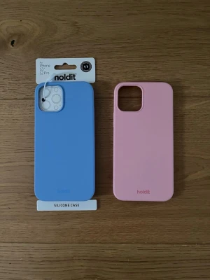 Blått och rosa mobilskal Holdit - Två snygga mobilskal från Holdit, ett i ljusblått och ett i ljusrosa. Båda är gjorda i mjukt silikon och passar iPhone 12/12 Pro. Skalen har en stilren design med öppning för kamera och logotypen diskret längst ner. Helt oanvända då de köptes i fel storlek! Ett för 130 kr, båda för 200 kr