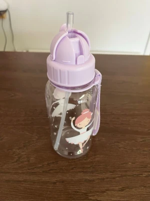 Vattenflaska med ballerina och sugrör - Söt vattenflaska i transparent plast med motiv av en dansande ballerina och lila lock. Flaskan har ett praktiskt sugrör och ett lila band för enkel bärning. Perfekt för att ta med till träning eller utflykter.