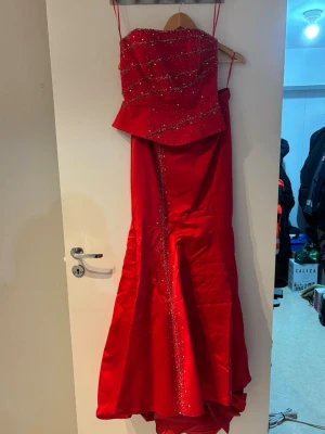 Röd långklänning med paljetter - Elegant röd långklänning med inga axelband och glittrande paljettdetaljer över byst och längs kjolen. Klänningen har en figurnära passform upptill och en lätt utsvängd kjol. Klänningen är också i två delar. Perfekt för bal eller festliga tillfällen.