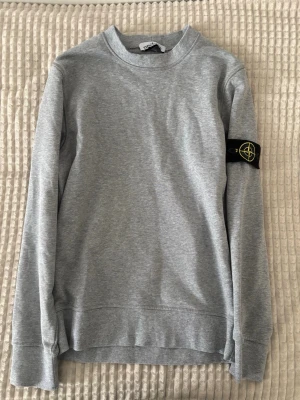 Grå sweatshirt från Stone Island - Snygg grå sweatshirt från Stone Island med klassisk rund hals och ikonisk svart patch med gul logga på vänster ärm. Tröjan har långa ärmar och är tillverkad i mjukt bomullsmaterial. Perfekt för en avslappnad och trendig stil.