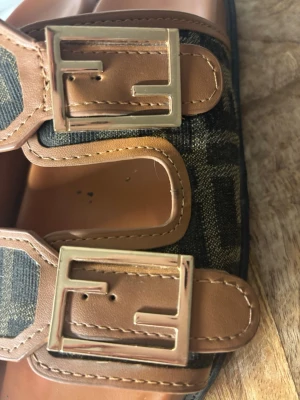 F**di sandaler - Snygga sandaler med klassisk FF-logga i brun och brunt tyg. Remmar i brunt läder med stora guldfärgade spännen. Mjuka innersulor i brunt och svart sula. Perfekta för en trendig och lyxig look. Använda men fortfarande jätte fina 