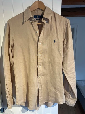 Beige linneskjorta från Ralph Lauren - Fin skjorta från Ralph Laurens linne skjorta kollektion i högkvalitativt linne. Skjortan har ”button-down” knappar till kragen och är i storlek S som jag skulle säga är normal i storleken och passar någon med skjortstorlek 37 eller 38. Använd men inga tecken på användning eller skador.