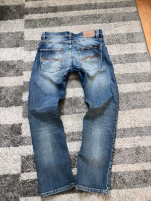 Nudie Jeans med slitningar - Säljer ett par klassiska blå jeans från Nudie Jeans med snyggt slitna detaljer och kontrastsömmar. Jeansen har raka ben, fem fickor och läderpatch bak i midjan. Perfekta för en avslappnad och trendig stil.