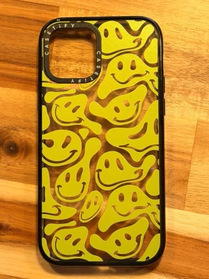Casetify skal smiley-mönster svart kant iPhone 12 Pro - Unikt mobilskal från Casetify med svart ram och transparent baksida dekorerad med gula och vita smiley-ansikten i ett lekfullt mönster. Skalet är gjort i tålig plast och har öppning för kamera. Perfekt för dig som vill ha ett skal som sticker ut men skyddar mobilen.