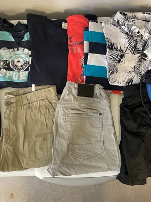 Beige shorts med knapp och fickor - Klädpaket pojke storlek 158-170  12 plagg Snygga beige shorts med knappstängning och sidfickor. Klassisk design med bälteshällor och normal passform. Perfekta för varma dagar och enkla att matcha med olika toppar.