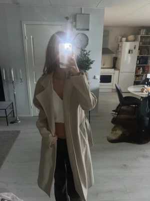 Beige lång kappa med bälte - Snygg beige kappa med bälte i midjan och breda slag. Lång modell som ger en stilren och trendig look. Perfekt för dig som vill ha en klassisk och enkel kappa till höst och vår. Kappan har en loose passform och är lätt att matcha med olika outfits.
