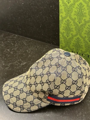 Beige keps från Gucci med GG-mönster - Snygg beige keps från Gucci med klassiskt GG-mönster i mörkblått. Kepsen har en böjd skärm och en ikonisk röd-blå rand på sidan. Materialet är canvas med brun insida och justerbar rem baktill. Perfekt för dig som vill ha en exklusiv accessoar.