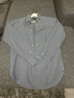 Blårutig skjorta från Polo Ralph Lauren - Säljer en blårutig skjorta från Polo Ralph Lauren i slim fit-modell. Skjortan har klassisk krage, långa ärmar och knappar framtill. Materialet är mjuk bomull och det finns en liten rosa logga broderad på bröstet. Perfekt för dig som gillar stilrena och tidlösa plagg.
