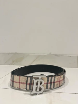 Burberry rutigt bälte med B-spänne - Snyggt bälte från Burberry med klassiskt rutigt mönster i beige, svart och rött. Bältet har ett stort silvrigt B-spänne framtill och är gjort i skinn. Perfekt accessoar för att lyfta din outfit.