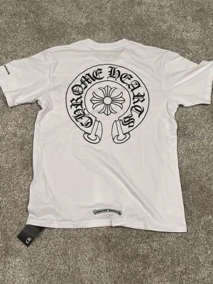 Vit Chrome Hearts t-shirt med tryck - Snygg vit t-shirt från Chrome Hearts med stort svart tryck på ryggen och logga nertill. Klassisk passform med rund hals och korta ärmar. Perfekt för dig som gillar streetwear och vill sticka ut med en ikonisk design.