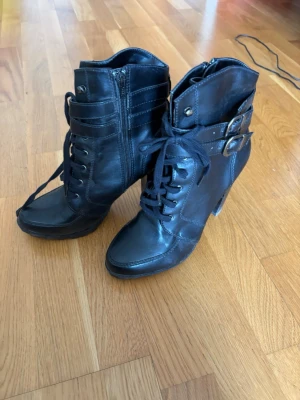 Svarta stövletter med snörning  - Snygga svarta stövletter i skinnimitation med snörning framtill, dragkedja på sidan och två dekorativa spännen vid ankeln. Klackarna är höga och ger en cool look. Perfekta för dig som vill ha en edgy stil.