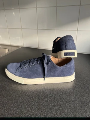 Blå mockasneakers från Polo Ralph Lauren - Snygga blå sneakers i mocka från Polo Ralph Lauren med klassisk låg profil och vit sula. Skorna har matchande blå skosnören och diskret Polo-logga på hälen. Perfekta för en stilren och avslappnad look. Modell: Jermaine! Tillverkade i Mocka/Läder! (Äkta) Skinn även inuti! Färg: Blå Storlek 43, UK: 9, US: 10 Innermått ca: 29,2 cm. Skorna är i mycket Fint skick!