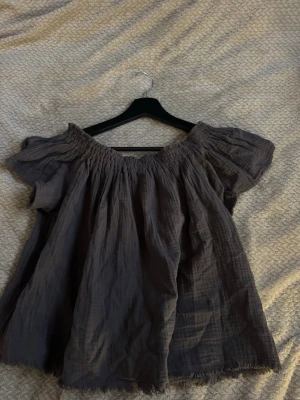 Grå offshoulder blus i bomull - Säljer en luftig grå blus med offshoulder-modell och korta ärmar. Toppen har en rynkad resår vid halsen och är tillverkad i ett mjukt bomullstyg. Perfekt för dig som gillar en avslappnad och trendig stil.