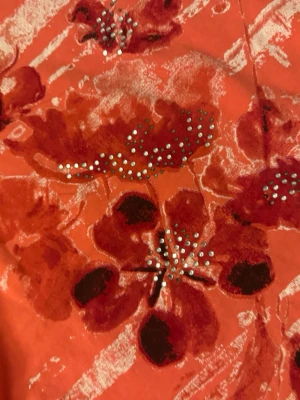 Orange t-shirt med blommor och strass - Orange t-shirt med röda och mörkröda blommor, dekorerad med små strasstenar på framsidan. T-shirten har rund halsringning och korta ärmar. Materialet ser ut att vara bomull eller bomullsblandning. Perfekt för dig som gillar färg och detaljer!