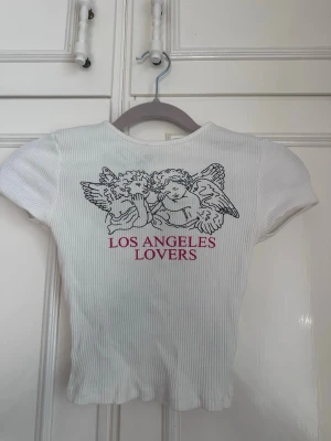 Vit ribbad topp med änglar - Vit ribbad kortärmad topp med tryck av två änglar och texten 'LOS ANGELES LOVERS' i rött på bröstet. Figurnära passform och rund halsringning. Perfekt för en trendig och avslappnad stil. Har en liiiiten fläck under armhålorna men går nog bort med galltvål i tvätten! Super snygg sommar topp💕