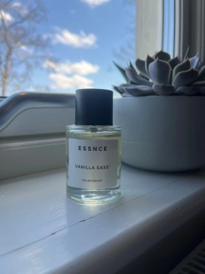 ESSNCE Vanilla Sass Eau de Parfum - Parfymen Vanilla Sass från ESSNCE kommer i en stilren, rund glasflaska med svart lock. Doften har tydliga inslag av vanilj och flaskan har en minimalistisk vit etikett. Perfekt för dig som gillar söta och fräscha dofter.