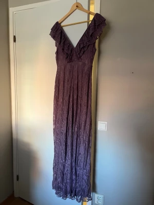 Lång lila spetsklänning med volang - Långklänning i lila spets med djup v-ringning och volangdetaljer längs axlarna. Klänningen har ärmlös design och ett elegant, figurnära fall. Klänningen har använts endast en gång och är då i ett mycket fint skick.😁