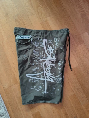 Gröna Billabong shorts med tryck - Säljer ett par gröna shorts från Billabong med stort vitt grafiskt tryck och logga på sidan. Shortsen har en ficka med kardborre och snörning i midjan. Perfekta för stranden eller poolen, tillverkade i snabbtorkande material.kan ha vanligt med 