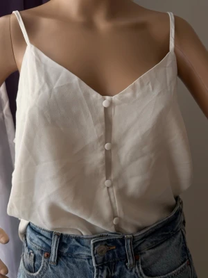 Vit linnetopp med knappar - Snygg vit linnetopp med smala axelband och dekorativa knappar framtill. Toppen har v-ringning och är tillverkad i ett lätt och luftigt material, perfekt för varma dagar. Enkel och stilren design som passar till jeans eller kjol.