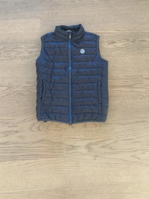 Mörkblå dunväst från North Sails  - Snygg mörkblå dunväst från North Sails med quiltad design och hög krage. Västen har en dragkedja framtill och två sidofickor. Insidan är blå med ljusa detaljer och märkets logotyp syns både på bröstet och insidan. Perfekt för lager-på-lager under kalla dagar.