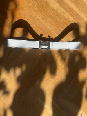 Vitt skärp med silverfärgat H-spänne - Stilrent vitt bälte med ett stort, silverfärgat H-format spänne. Bältet har en slät yta och är tillverkat i skinn, vilket ger en lyxig känsla. Perfekt accessoar för att lyfta din outfit med en modern touch.