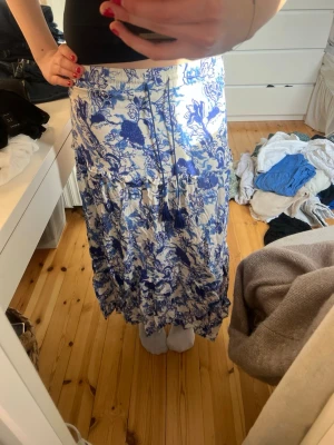 Blåvit långkjol med blommigt mönster - Säljer en flowy långkjol i vitt med blått blommigt mönster. Kjolen har resår i midjan och snörning med tofsar framtill. Perfekt för dig som gillar bohemisk stil och vill ha något luftigt till sommaren.