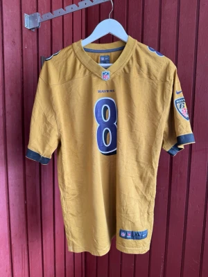 Gul NFL Ravens tröja Jackson #8 - Snygg gul NFL-tröja från Nike med Ravens-logga och nummer 8 på bröstet och ryggen. JACKSON står tryckt på ryggen. Tröjan har korta ärmar och svarta detaljer vid ärmslut och axlar. Perfekt för dig som gillar amerikansk fotboll och vill sticka ut. Ungdoms storlek men passar även Dam S-M