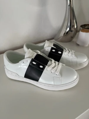 Valentino Garavani vita sneakers - Snygga vita sneakers från Valentino Garavani med bred svart läderdetalj över mitten och vita skosnören. Klassisk låg modell med gummisula och diskret logga på plösen. Perfekta för dig som gillar stilrena och exklusiva sneakers.
