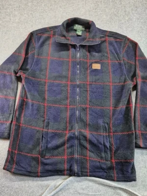 Mörkblå Lauren by Ralph Lauren fleecejacka med rött rutmönster  - En Fleece jacka från Lauren by Ralph Lauren! Se bröst och längdmått på sista bilderna så du vet den passar dig perfekt! Har även med ärmlängd och då är det mätt från övre axelsöm ned till.  hel dragkedja och rött rutmönster. Jackan har ståkrage, två fickor framtill och en liten läderpatch med logga på bröstet. Perfekt för kyliga dagar och enkel att matcha med jeans eller mjukisbyxor. Storlek M( men rätt så stor så mer L) Pris kan diskuteras. 
