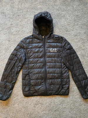 Svart pufferjacka från EA7 Emporio Armani - Snygg svart pufferjacka från EA7 Emporio Armani med huva och dragkedja framtill. Jackan har quiltad design, två sidofickor och logga på bröstet. Perfekt för kalla dagar när du vill hålla stilen och värmen.