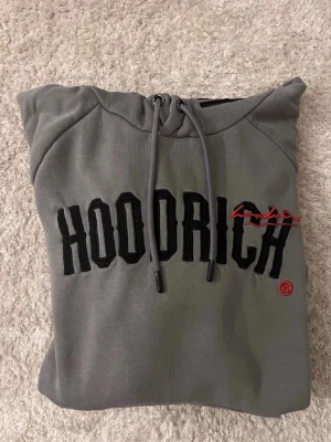 Grå hoodie från Hoodrich - Snygg grå hoodie från Hoodrich med svart broderad logga och röd detalj på bröstet. Klassisk känguruficka framtill, dragsko i huvan och ribbade muddar. Perfekt för en streetwear-look och riktigt skön att ha på sig.
