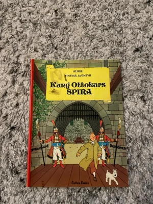 Tintins äventyr: Kung Ottokars spira - Säljer seriealbumet 'Kung Ottokars spira' av Hergé ur Tintins äventyr. Boken har hårda pärmar med färgglad framsida där Tintin och Milou syns vid en borgport. Perfekt för dig som gillar klassiska serier och äventyr.