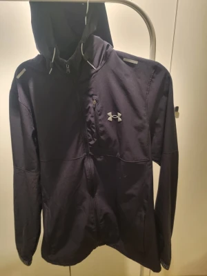 Svart vindjacka från Under Armour - Svart vindjacka från Under Armour med huva och dragkedja framtill. Jackan har en bröstficka med dragkedja och detaljer på axlarna. Fast pris 550 kr.