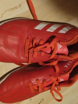 Röda Adidas Copa fotbollsskor - Säljer ett par röda Adidas Copa fotbollsskor med klassiska vita ränder på sidan. Skorna har snörning och är tillverkade i syntetmaterial med en sportig design. Perfekta för dig som vill sticka ut på planen och köra hårt i matchen.