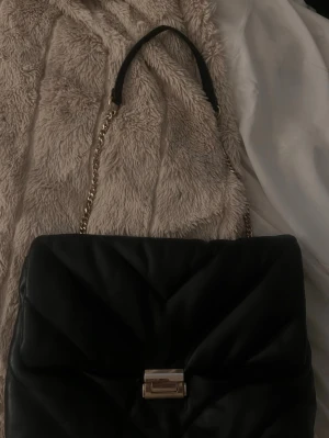 Svart quiltad handväska med guldkedja - Snygg svart quiltad handväska med guldfärgad kedjerem och elegant guldfärgat spänne framtill. Väskan har en mjuk, puffig look och är tillverkad i konstläder. Perfekt accessoar för att lyfta din outfit.