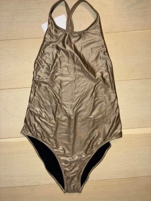 Guldig baddräkt med korsade band - Snygg guldig baddräkt från Filippa K med glansig finish och korsade axelband i ryggen. Baddräkten har en hög skärning och djup rygg, vilket ger en trendig och sportig look. Perfekt för stranden eller poolen.