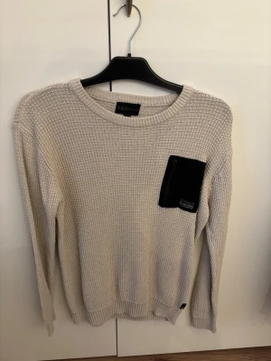 Beige stickad tröja med svart ficka - Snygg beige stickad tröja med rund halsringning och en svart bröstficka som cool detalj. Tröjan har lång ärm och ribbade muddar vid ärmslut och nederkant. Perfekt för dig som gillar stilrena plagg med en twist.