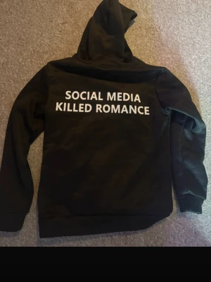 Svart hoodie med text på ryggen - Säljer en svart hoodie med huva och trycket 'SOCIAL MEDIA KILLED ROMANCE' i vitt på ryggen. Hoodien har klassisk passform, ribbade muddar och är gjord i mjukt material. Perfekt för dig som gillar statement-plagg och vill sticka ut. Aldrig använd, nypris 800kr. 