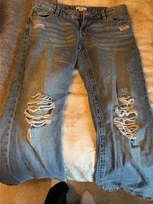 Blå slitna jeans från Gina Tricot - Säljer ett par blå jeans från Gina Tricot med stora slitningar och hål på båda knäna. Jeansen har klassisk femficksmodell, dragkedja och knapp framtill. Perfekta för en avslappnad och trendig look.