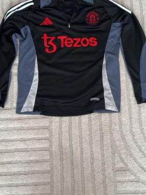 Manchester United träningströja Adidas - Svart och grå långärmad träningströja från Adidas med Manchester United-logga och röd Tezos-sponsor på bröstet. Tröjan har dragkedja vid halsen och vita detaljer på axlarna. Perfekt för fotbollsträning eller sportiga dagar.