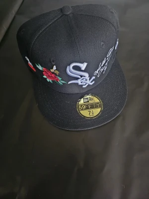 Svart White Sox keps New Era 59FIFTY - Snygg svart keps från New Era 59FIFTY med White Sox-logga framtill, broderade blommor och detaljer på sidorna samt patch med '75 Years'. Platt skärm och klassisk passform. Perfekt för dig som gillar streetwear och sportiga accessoarer.