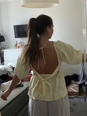 Gul blus med öppen rygg och knyt - Söt gul blus med puffärmar, öppen rygg och knyt både i nacken och baktill i midjan. Luftig och perfekt för varma dagar, med en avslappnad men ändå trendig vibe. Passar dig som gillar detaljer och vill sticka ut lite extra.