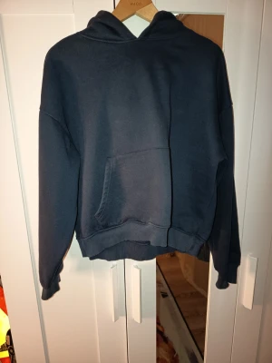 Mörkblå hoodie med magficka - En basic mörkblå hoodie med huva och stor magficka framtill. Tröjan har ribbade muddar vid ärmslut och nederkant, samt en avslappnad passform. Perfekt för en chill och avslappnad stil. Materialet känns mjukt och bekvämt, troligtvis bomullsblandning.