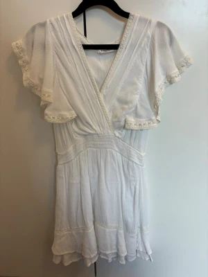 Vit kort klänning med volangärmar - Söt vit klänning med omlottliv, volangärmar och spetsdetaljer. Klänningen har en lätt rynkad midja och faller mjukt med volanger nertill. Perfekt för varma dagar och har en romantisk känsla tack vare spetsen och den luftiga designen.