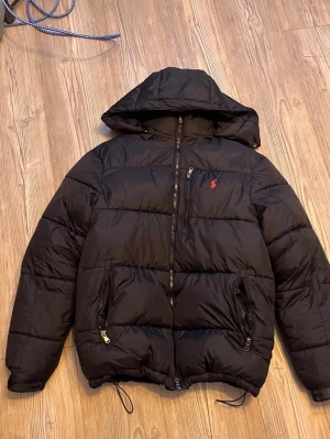 Svart pufferjacka från Polo Ralph Lauren - Svart pufferjacka med huva från Polo Ralph Lauren. Jackan har dragkedja framtill, två fickor med dragkedja och den klassiska röda Polo-loggan broderad på bröstet. Perfekt för kalla vinterdagar och riktigt skön att bära. Storlek small, pris kan diskuteras, hör av dig!