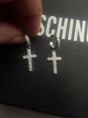 Silvriga kors örhängen med stenar - Snygga örhängen från Moschino i silverfärg med korsformade hängen täckta av glittrande stenar. Perfekta för dig som gillar statement-smycken och vill sticka ut med en trendig accessoar.