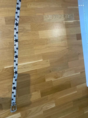Glittrigt bälte med stjärnmönster - Snyggt bälte med glittriga stenar och svartvita stjärnor. Bältet har en rektangulär spänne täckt av små stenar som ger extra bling. Perfekt för att lyfta vilken outfit som helst med en cool och unik touch. (Slitet vid hålen)