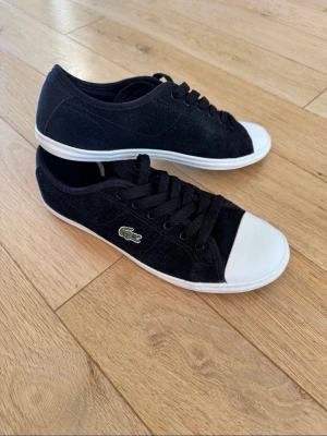 Svarta Lacoste sneakers med vit tå - Snygga svarta sneakers från Lacoste med klassisk vit gummitå och vit sula. Skorna har snörning och den ikoniska gröna krokodilloggan på sidan. Ovandelen är i mockaliknande material som ger en stilren och sportig look. Storlek 38, mycket bra skick. Pris kan diskuteras, hör av dig!