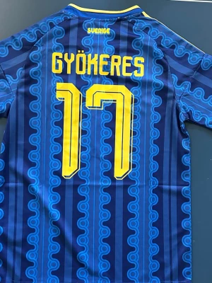 Sverige fotbollströja Gyökeres #17 - Ny Sveriges landslagströja inför VM 2026 och Gyökeres nummer 17 i gult. Tröjan har korta ärmar, V-ringning och gula detaljer på med unikt blått mönster. Storlek S