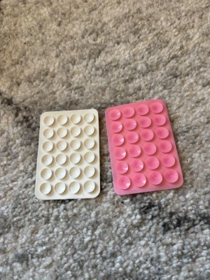 Två mobilgrepp Octopus octobuddy silikon - Säljer två rektangulära pop it fidget-leksaker i silikon, en i vitt och en i rosa. Varje leksak har flera bubblor att trycka på och är perfekt för att sysselsätta händerna eller stressa ner.